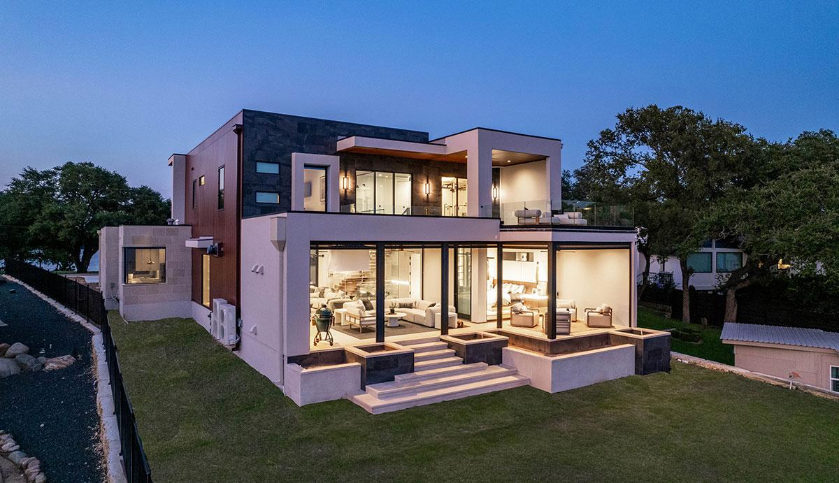 Evening view of a modern luxury lakehouse on Lake LBJ designed by Zbranek & Holt Custom Homes, showcasing expansive glass walls, a spacious terrace with seating, and soft interior lighting that highlights the home’s elegant design and seamless indoor-outdoor living spaces.